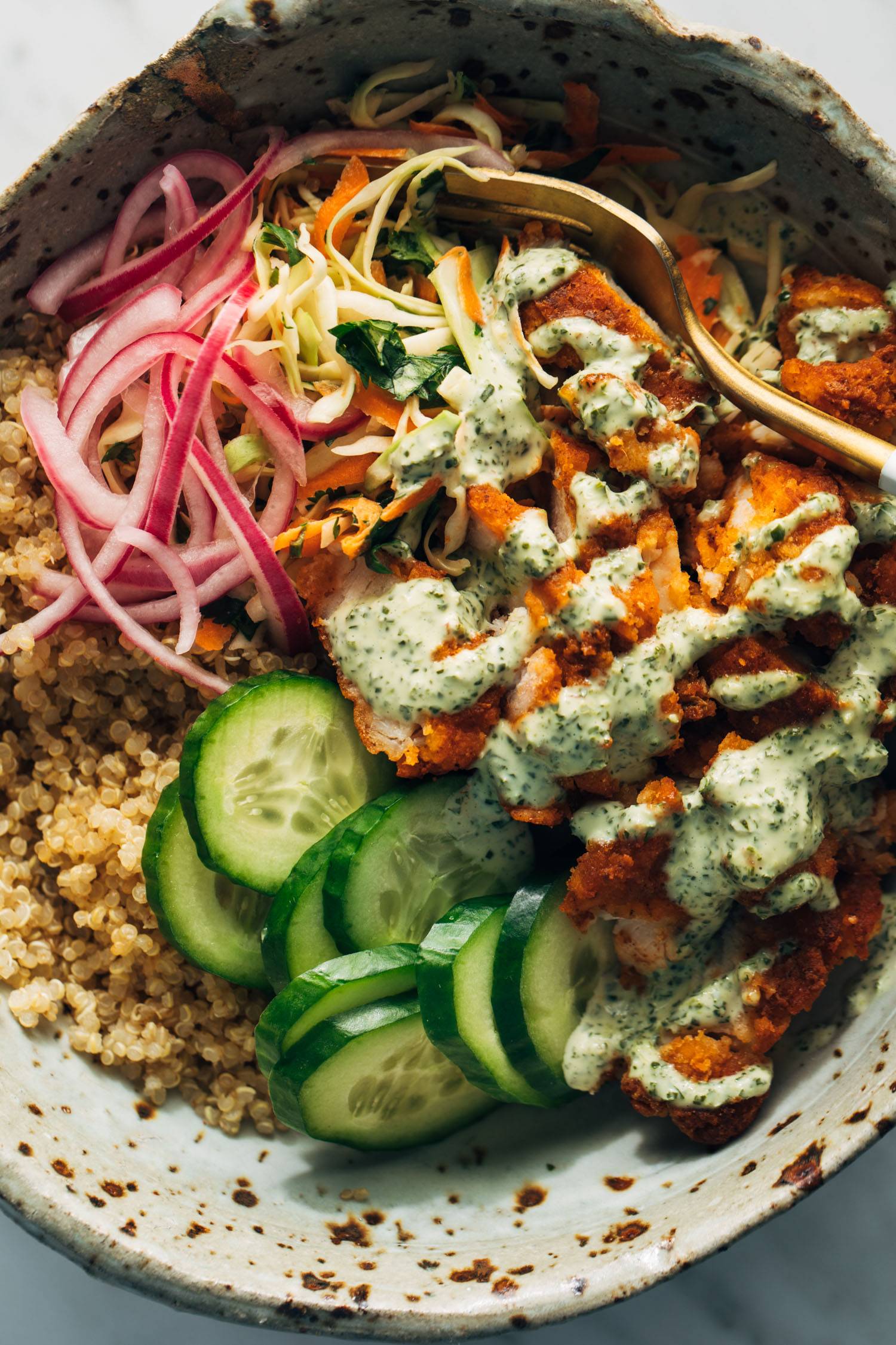 Crispy chicken, quinoa, cucumbers, and pickled onions in a bowl with mint sauce and a fork