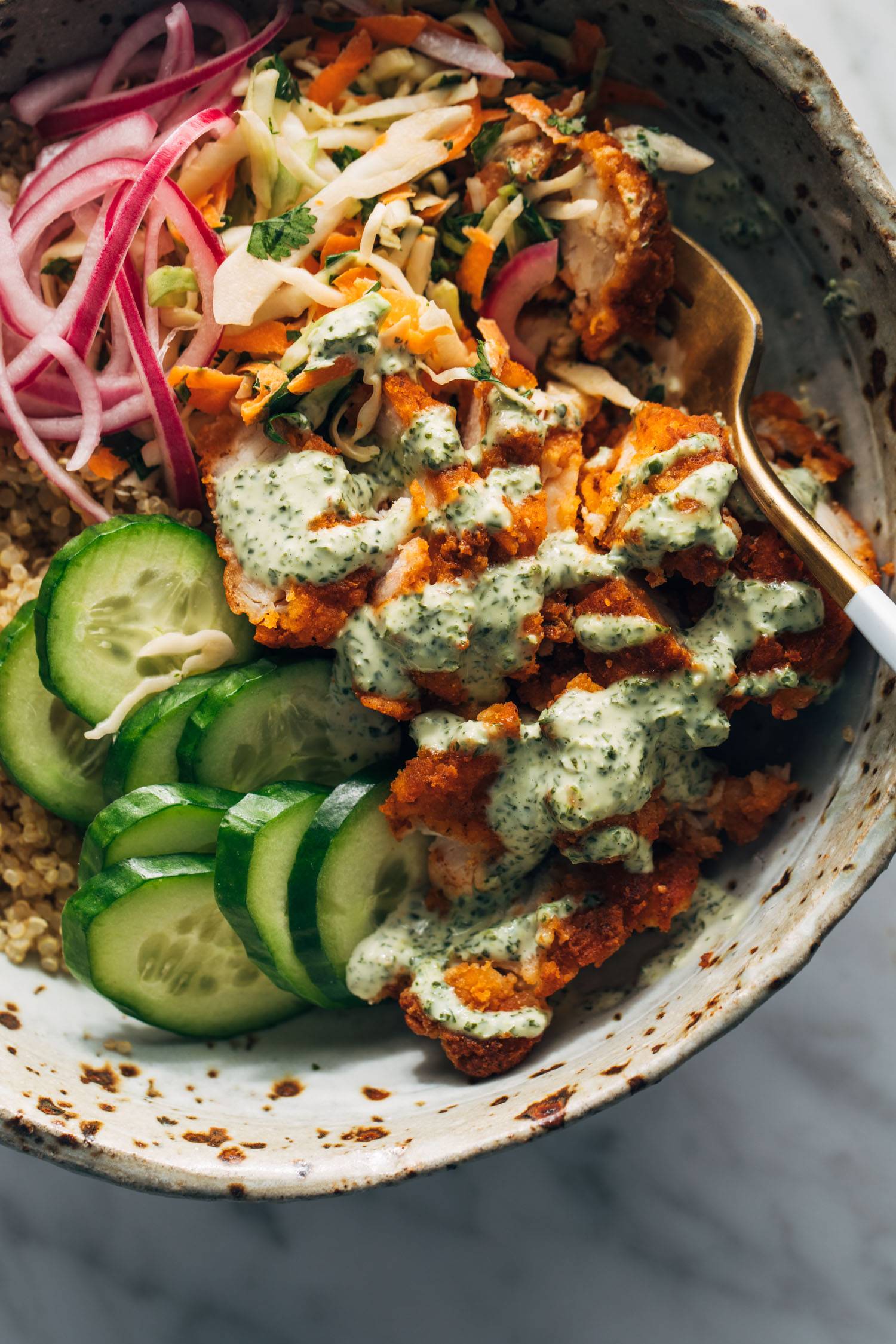 Crispy chicken, cucumbers, quinoa, and pickled red onions in a bowl with mint sauce and a fork