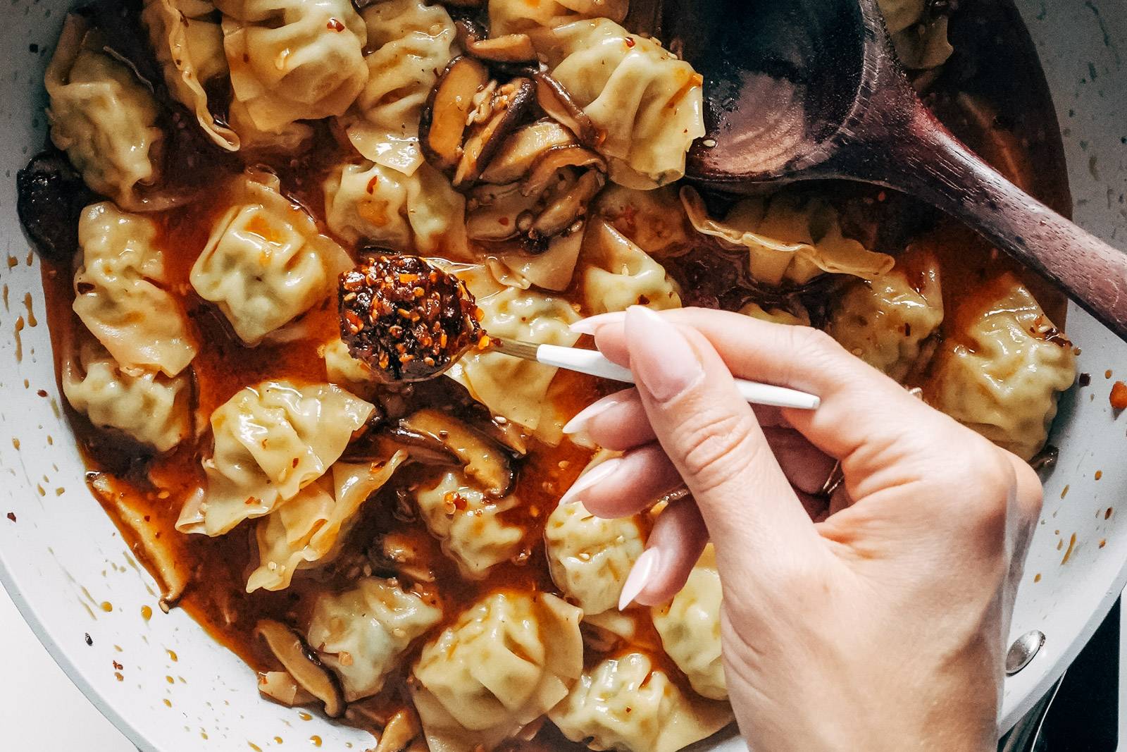 White hand adding chili oil to chicken wontons in a pan.