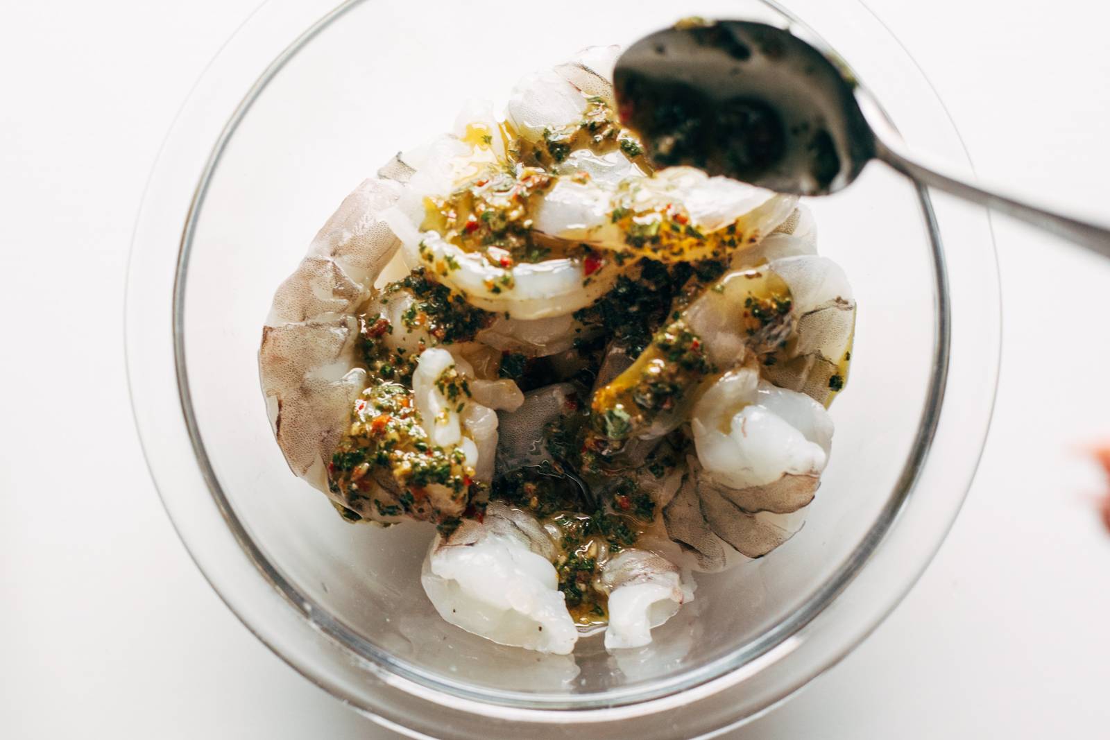 Spooning chimichurri sauce onto raw shrimp in a bowl.