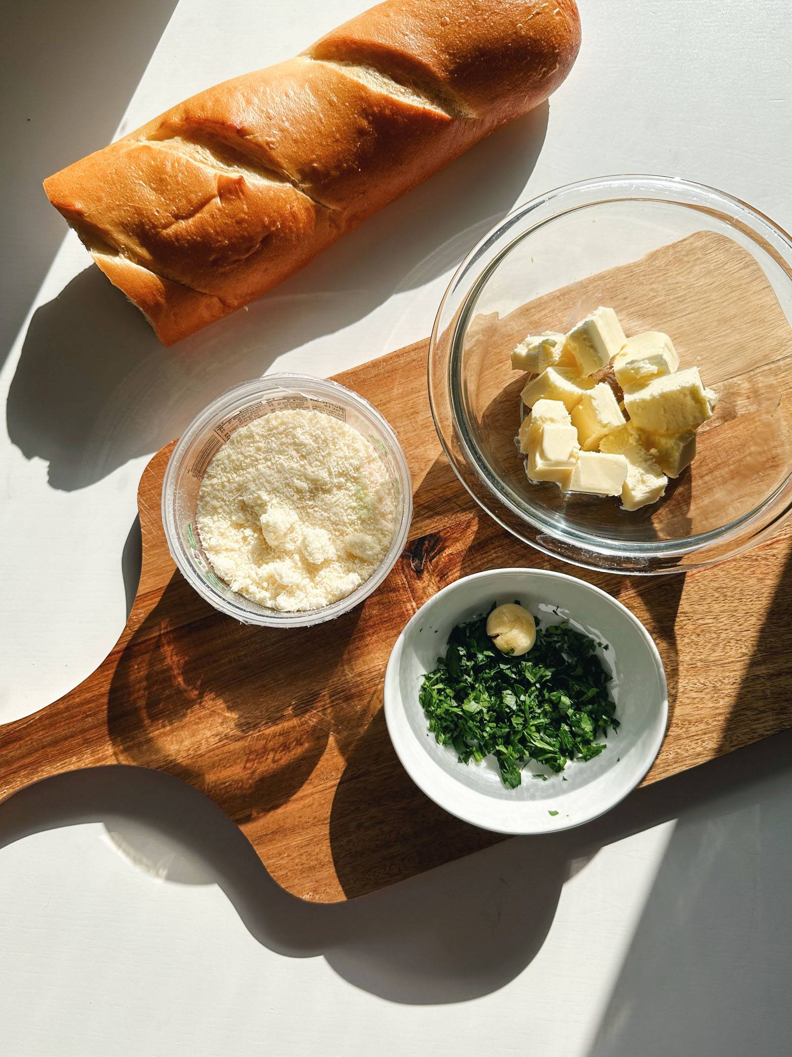 Ingredients for garlic bread - a baguette, parmesan cheese, butter, garlic, and parsley.