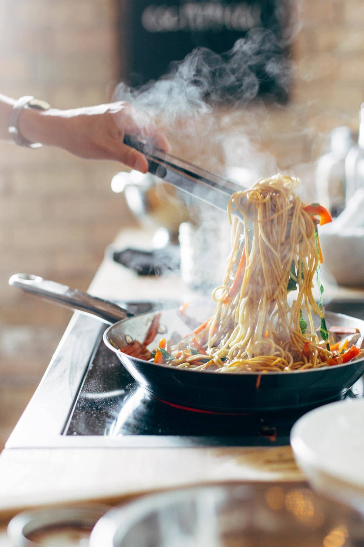 White hand grabbing noodles in a pan with a tong. 